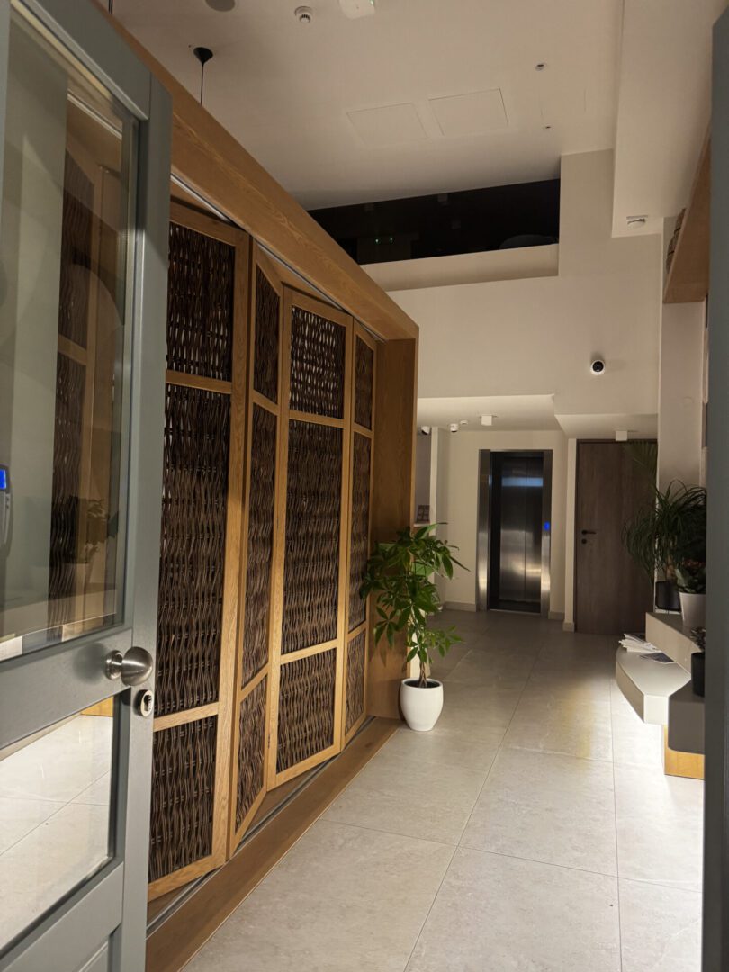 Open wooden door leading to a bright, modern hallway with potted plants.