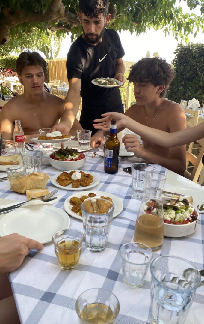 Two shirtless young men enjoying a meal outdoors with various dishes and drinks.