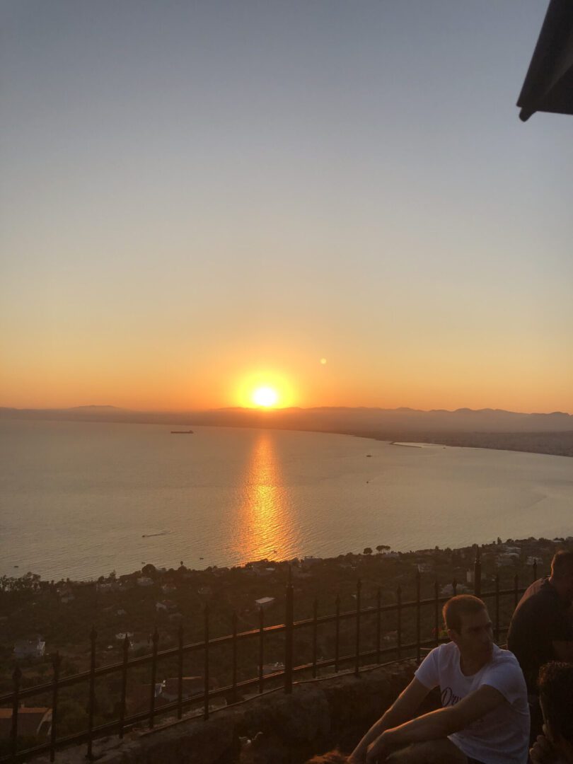Sunset over a calm sea with silhouetted land and people in the foreground.