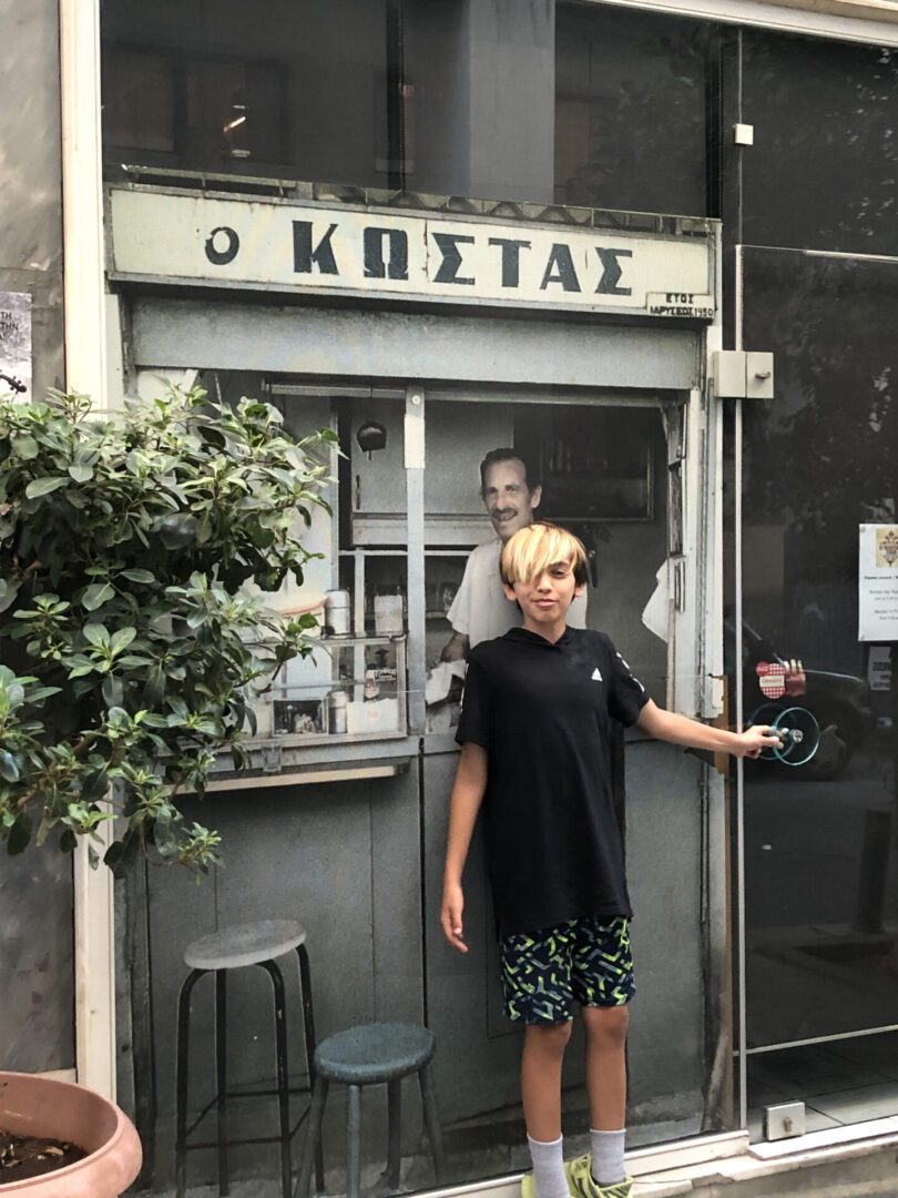 Person entering a small, rustic café named "Ο ΚΩΣΤΑΣ".