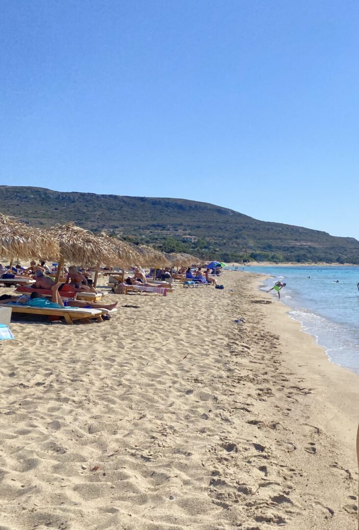 Crowded beach with sun loungers and clear blue water under a sunny sky.